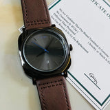 Watch with brown leather strap and black dial on a white surface with a certificate of authenticity.